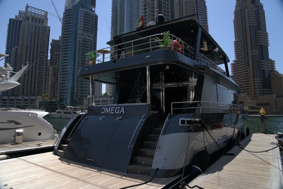The stern of a modern, dark grey luxury yacht named 'OMEGA' docked at a marina, with the towering skyscrapers of Dubai Marina in the background.