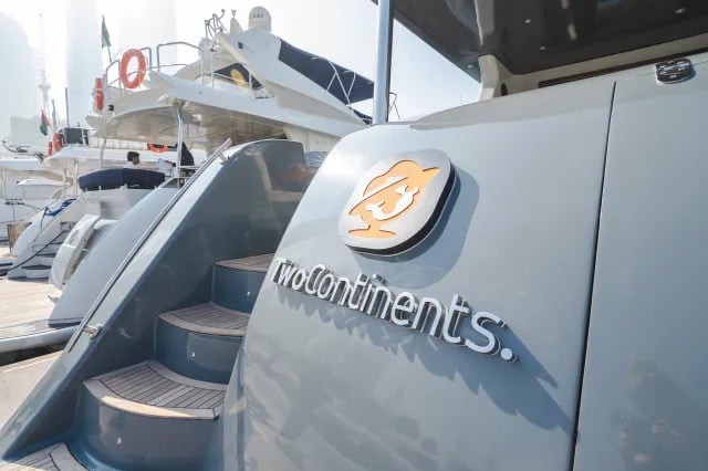 The stern of a modern, dark grey luxury yacht named 'OMEGA' docked at a marina, with the towering skyscrapers of Dubai Marina in the background.