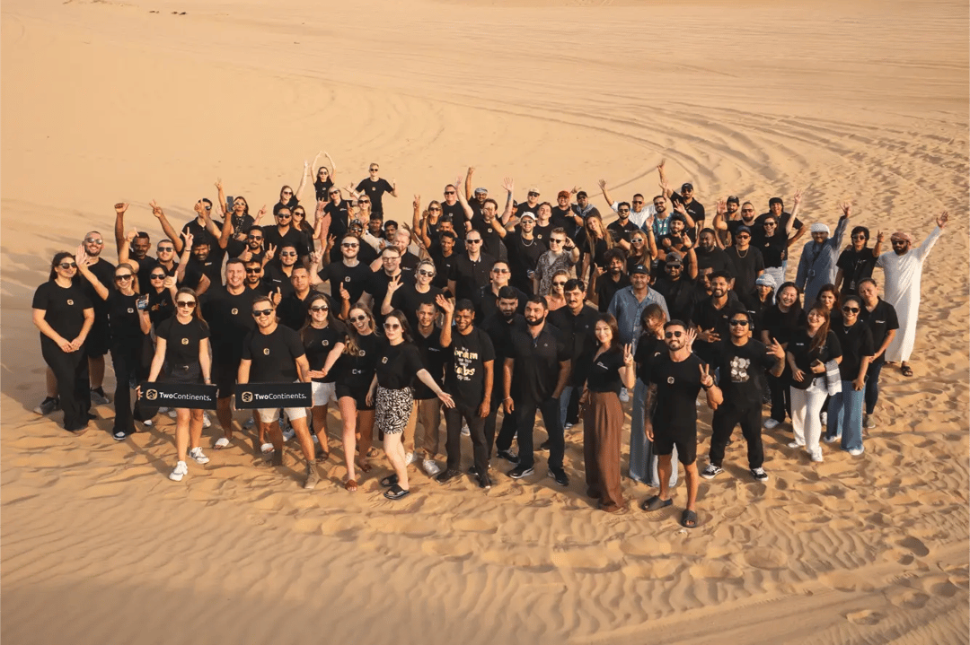 A large group from the TwoContinents team posing for a photo on a vast sand dune, smiling and waving at the camera.