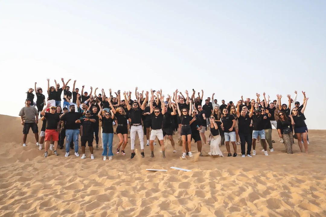The TwoContinents team celebrating together in the desert, all jumping simultaneously with their arms raised in excitement against a clear sky.