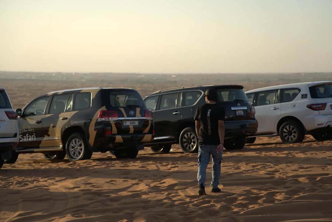 A man in a TwoContinents t-shirt walks across the sand towards a line of safari SUVs parked in the desert during a warm, hazy sunset.