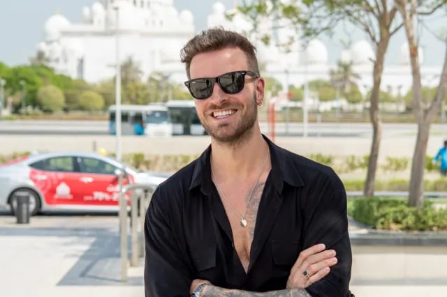 A man in a TwoContinents t-shirt walks across the sand towards a line of safari SUVs parked in the desert during a warm, hazy sunset.